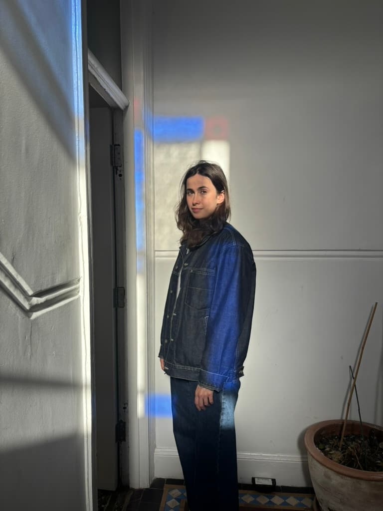 Ana Dunne Sequi in a denim jacket, looking at the camera in a sunlit white hallway with blue window light on the wall